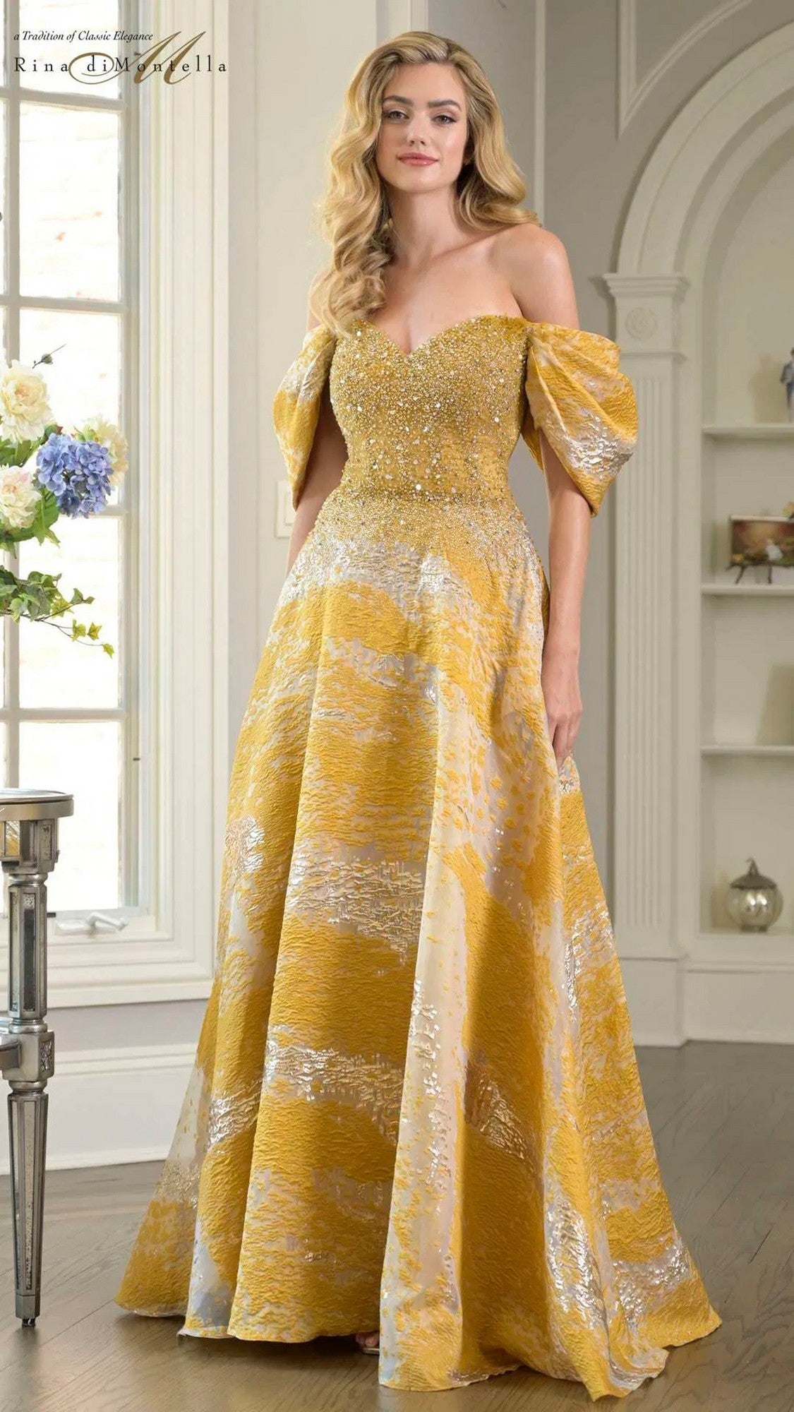 A woman stands indoors wearing the gold Rina Di Montella RD3405 long formal dress, featuring off-the-shoulder puff sleeves, a beaded bodice, and an elegant a-line organza skirt.